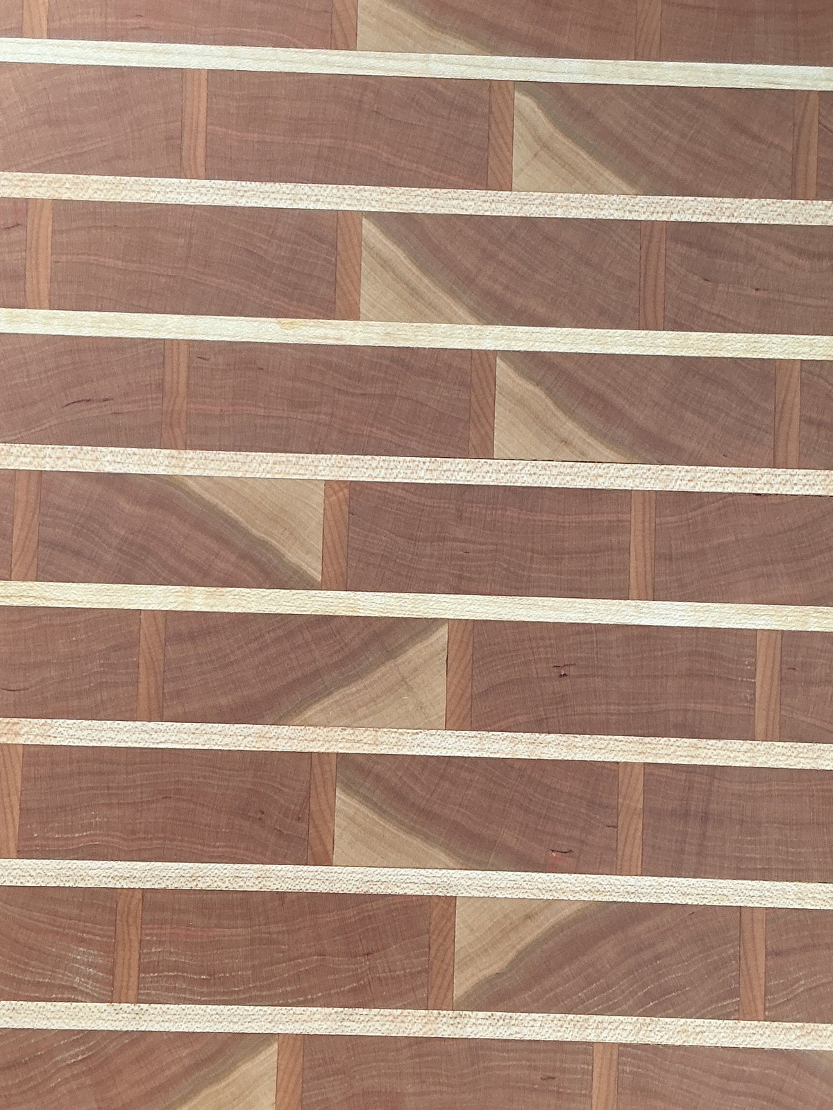 A rectangular wooden cutting board with a striped and geometric inlay pattern made of different wood tones, shown from above on a table surrounded by crafting supplies.